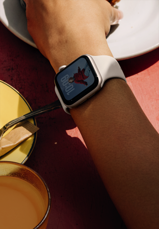 Braccio di una persona che fa colazione, Apple Watch SE 3 al polso, cassa in alluminio color galassia, display always-on, ritratto di una persona sul quadrante, cinturino Sport color galassia